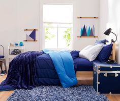 a bed with blue sheets and pillows in a white room next to a large window