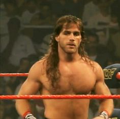 a man with long hair and no shirt standing in front of a roped off wrestling ring