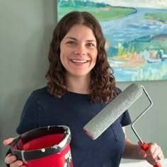 a woman holding a paint roller and a red bucket in her right hand while smiling at the camera