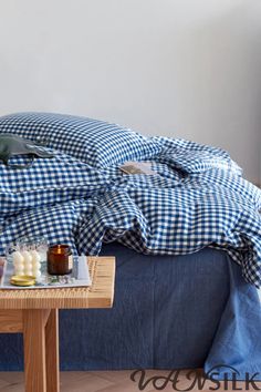 an unmade bed with blue and white checkered comforter on it, next to a small wooden table