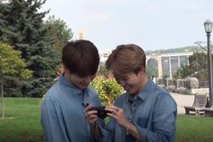 two young men standing next to each other in front of trees and grass, looking at their cell phones