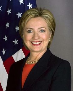 a woman standing in front of an american flag and wearing a black suit with gold necklace