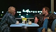 a man and woman sitting at a table in front of a window with the city lights behind them