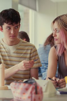 two people sitting at a table with food and pens in their hands while looking at something on the phone