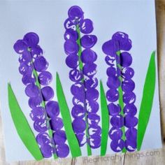 purple flowers are painted on white paper with green stems in the center and behind them is a piece of wood