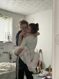 two women hug each other while taking a selfie in a bedroom with an unmade bed