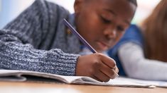 a young boy writing on a book while sitting at a desk with a pencil in his hand