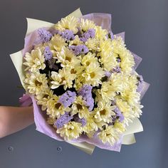 a bouquet of yellow and purple flowers is held by someone's hand in front of a gray wall