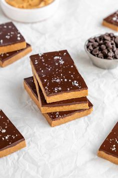 several pieces of chocolate and peanut butter bars on white paper with bowls of cookies in the background