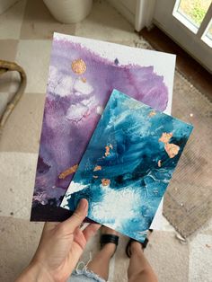 someone is holding two paintings in their hands, one has blue and purple paint on it