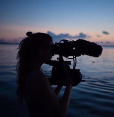 a woman holding a camera in front of the ocean at dusk with clouds behind her