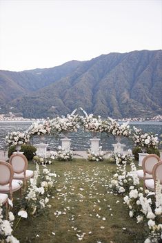 an outdoor ceremony setup with chairs and flowers on the grass by the water in front of mountains