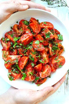 two hands holding a white bowl filled with sliced tomatoes and parsley on the side