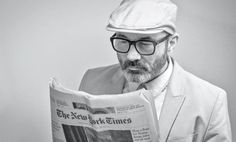 a man reading a newspaper while wearing glasses and a hat on top of his head