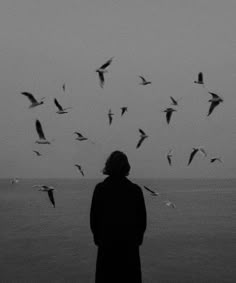 a person standing in front of a flock of birds flying over the ocean on a cloudy day