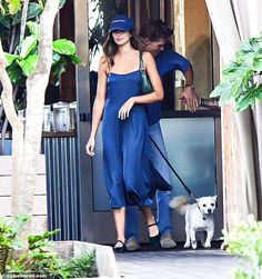 a woman in a blue jumpsuit walking her dog through an open door with another person