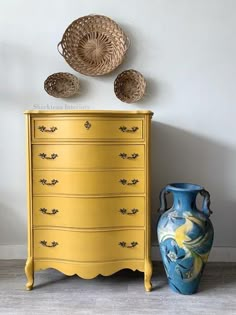 a yellow dresser next to a blue vase and wicker basket on top of it