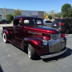 an old red truck parked in a parking lot next to other cars on the street