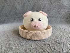 a crocheted pig sitting in a bowl on top of a gray blanket with eyes wide open