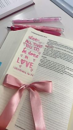 an open book with pink ribbon and writing on it next to some pens, scissors and a pen