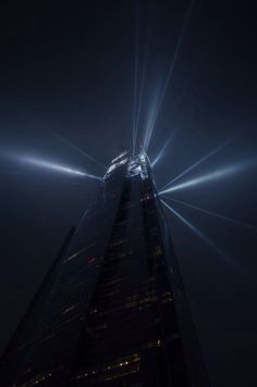 the top of a tall building lit up at night with bright beams in the sky