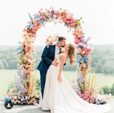 a bride and groom kissing in front of an archway with colorful flowers on the side