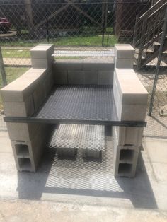 an outdoor bench made out of cinder blocks and concrete blocks, sitting on the sidewalk in front of a chain link fence