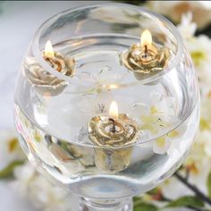 three lit candles in a glass bowl with white flowers behind it on a table top