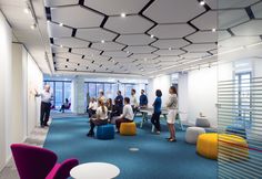 an office filled with people sitting and standing in front of large glass windows on the ceiling