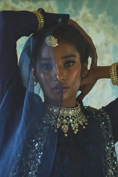 a woman in a black dress with gold jewelry on her neck and hands behind her head