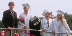 four people in graduation gowns and caps standing on a railing with ribbon around them