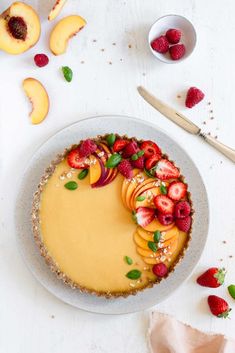 a cheesecake topped with sliced peaches and strawberries on a white table next to bowls of fruit