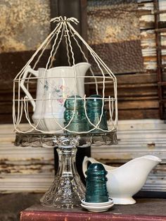 there is a bird cage with cups in it on top of a table next to a white pitcher