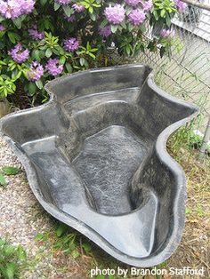 a large gray planter sitting on top of a gravel ground next to purple flowers