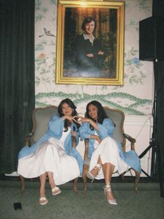 two women sitting on chairs in front of a wall with a painting behind them and one holding a camera