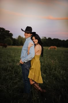 a man and woman standing together in a field with horses grazing behind them at sunset