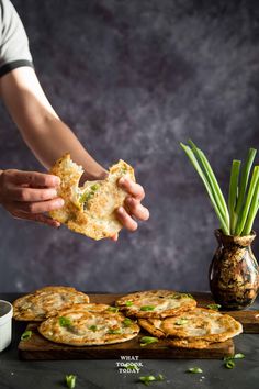 a person holding food in their hand near some other foods on a wooden board and a vase with green onions