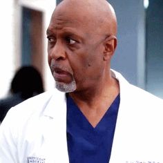 a bald man wearing a white lab coat and blue scrubs is looking at the camera