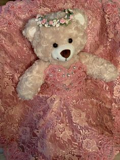 a teddy bear in a pink dress sitting on a bed with lace and flowers around its neck