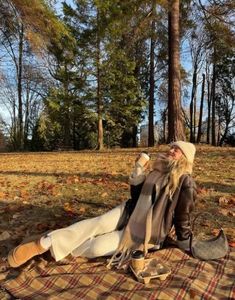 a woman sitting on top of a blanket next to a forest filled with trees and leaves