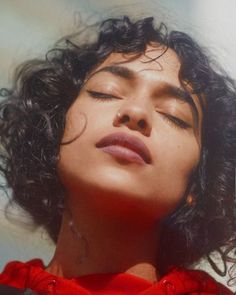 a woman with her eyes closed wearing a red shirt and black curly hair is looking up at the sky