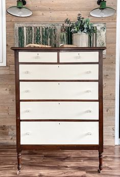 an old dresser with books and plants on top in front of a wooden wall behind it