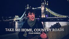 a young man is playing an acoustic guitar in front of the london bridge and slogan take me home, country roads