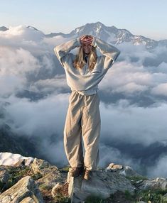 a woman standing on top of a mountain with her hands behind her head and clouds in the background