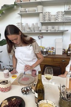 a woman in an apron preparing food on a table with plates and wine bottles behind her