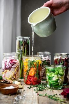 a person pours water into jars filled with different types of vegetables and herbs on a wooden table