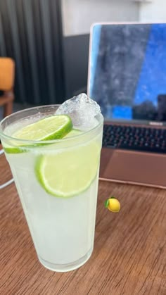 a glass filled with ice and lime sitting on top of a wooden table next to a laptop