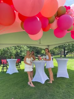 two women in white dresses standing under a tent with balloons hanging from it's ceiling