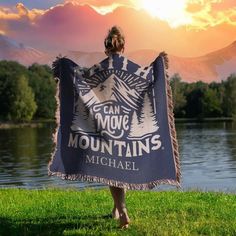 a woman is holding up a blanket that says can move mountains on it in front of a lake