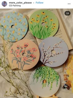 four different designs on paper with flowers and plants painted on them, sitting next to an open book